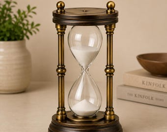 May include: An hourglass featuring a dark bronze-toned metal frame with a circular base and top, connected by four vertical pillars. The clear glass bulb contains white sand, with a portion of the sand currently resting in the bottom chamber.