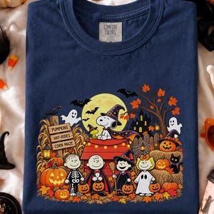 May include: A navy blue crew neck t-shirt featuring a graphic print of Snoopy and friends in Halloween costumes. The design includes a full moon, a red doghouse, pumpkins, and signs reading Pumpkins, Hay-Rides, and Corn Maze. The shirt has a Comfort Colors label.