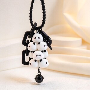May include: A black and white keyring featuring a stack of five small panda figures with pink cheeks. The charm includes a black silicone coil ring, a black plastic chain link, a black tag with the text "stuck with", and a small black bell.