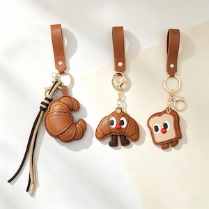 Peut inclure: Trois porte-clés en cuir marron avec des motifs sur le thème de la nourriture. L'un est un croissant simple avec un long pompon, un autre est un croissant personnage avec des yeux et un nez rouge, et le dernier est une tranche de pain.