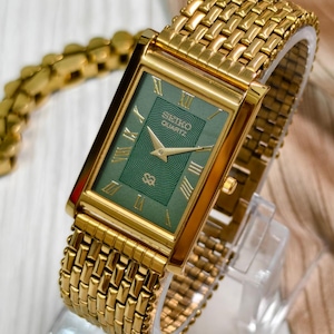 May include: A gold-tone rectangular wristwatch featuring a green dial with a textured center pattern, gold Roman numeral hour markers, and gold hands. The watch includes a matching gold-tone metal link bracelet and displays the text Seiko Quartz.