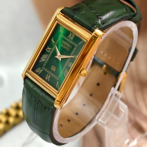 May include: A rectangular Seiko Quartz wristwatch featuring a gold-tone case and a vibrant emerald green dial with Roman numeral hour markers. The watch is paired with a dark green textured leather strap that has a glossy finish and gold buckle.