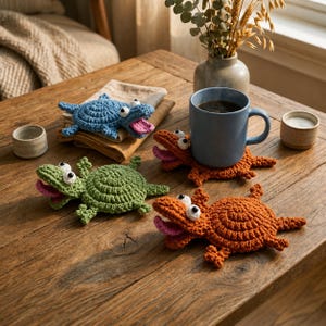 May include: Three handmade crochet coasters shaped like crocodiles in blue, green, and orange. Each coaster features a circular body, four small legs, a tail, and an open mouth with a pink tongue and two white eyes with black pupils.