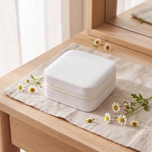 May include: A square, white jewelry box with a smooth, solid-colored exterior and a central zipper closure. The compact container features rounded corners and a clean, simple design, presented as a singular object for storage and organization.