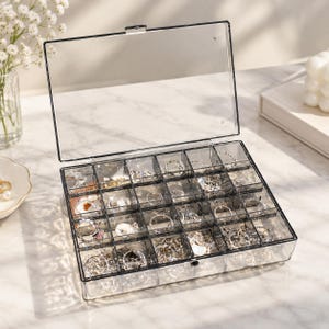 May include: A rectangular, clear acrylic jewelry organizer box with a hinged lid. The interior is divided into twenty individual square compartments, each containing various silver-toned rings, chains, and small metal jewelry pieces.