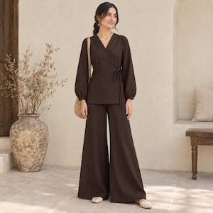 Può includere: Un completo due pezzi marrone scuro composto da un top a portafoglio a maniche lunghe con chiusura a fiocco laterale e pantaloni ampi coordinati. Il top presenta uno scollo a V e polsini arricciati, mentre i pantaloni hanno una linea fluida.