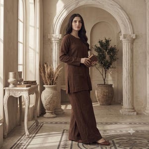 May include: A dark brown long-sleeved tunic top and matching wide-leg trousers set. The tunic features a round neckline with a vertical row of small buttons down the center front. The fabric appears to have a textured finish, and the outfit is worn with simple brown flat shoes.