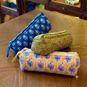 May include: Three cylindrical quilted fabric pencil cases with zippered closures. One is dark blue with a white leaf pattern, one is light green with a pink floral print, and one is yellow with a blue and pink floral pattern.