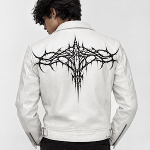 May include: A white leather jacket featuring a large, intricate black graphic print on the back. The design consists of sharp, thorn-like lines and pointed shapes arranged in a symmetrical, wing-like pattern. The jacket has a classic collar and zips.