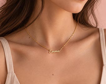 Puede incluir: Un collar de tono dorado con una cadena fina y un colgante central de placa con el nombre Emma escrito en letra cursiva. El collar descansa sobre el cuello, mostrando un acabado metálico pulido y una cadena de eslabones.