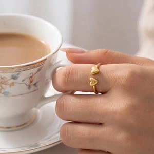 May include: A gold-toned open ring featuring two small, smooth heart-shaped charms at each end of the band. The ring is worn on a finger, positioned next to a white ceramic teacup decorated with a delicate floral pattern and gold trim.