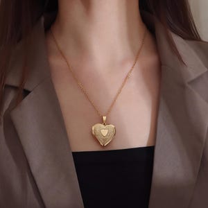 Puede incluir: Un collar de metal en tono dorado con un colgante de relicario en forma de corazón. El relicario presenta un intrincado patrón floral en relieve en su superficie con un corazón liso y sencillo en el centro. Cuelga de una cadena fina y delicada.
