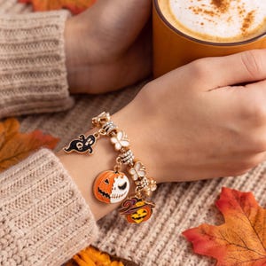 Op de afbeelding: Een goudkleurige bedelarmband met emaille bedels, waaronder een zwart spookje, een pompoen met een gespleten gezicht en een pompoen met een heksenhoed. De armband bevat kleine witte bloemvormige tussenstukjes en heldere kristallen op de schakels.