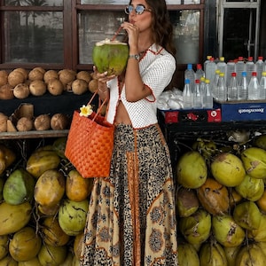 May include: A white crochet cardigan with orange trim, a long skirt featuring a black and tan floral pattern, and a bright orange woven tote bag. The outfit is paired with brown flip-flops and round sunglasses, while holding a green coconut.