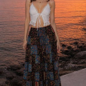 May include: A white sleeveless crop top with lace trim and a front tie closure paired with a long, tiered maxi skirt featuring a dark blue, black, and red floral paisley print. The outfit is completed with simple white strappy sandals.