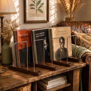 May include: A wooden vinyl record storage rack with three metal dividers holding several albums, including Miles Davis Sketches of Spain, John Coltrane A Love Supreme, and Bill Evans Trio Sunday at the Village Vanguard, displayed on a wooden table.