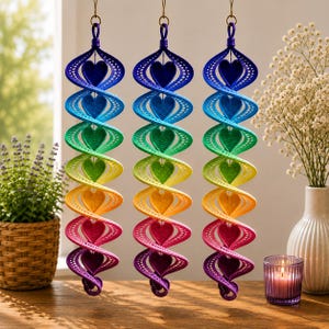 May include: Three hanging spiral ornaments featuring a vertical gradient of rainbow colors including blue, green, yellow, orange, and purple. Each ornament has a series of heart-shaped cutouts along the center and a textured, perforated surface.