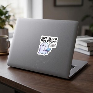 May include: A white vinyl sticker featuring a cartoon ghost holding a small laptop. The text reads 404: SLEEP NOT FOUND, Status: Critically low (Needs caffeine). The ghost has a tired expression, and the design includes a battery icon.