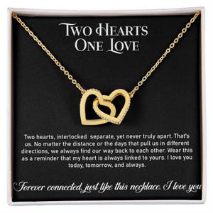 Pode incluir: Um colar de tom dourado com dois pingentes de coração entrelaçados, apresentado numa caixa de presente preta. A caixa exibe o texto Two Hearts One Love, seguido por uma mensagem sentimental e a frase Forever connected, just like this necklace.
