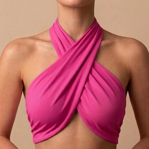 May include: A bright fuchsia halter top featuring a crisscross neckline design. The fabric wraps around the neck and across the chest, creating a gathered effect with soft folds. The garment is sleeveless and cropped, exposing the midriff area.