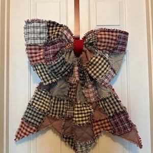 May include: A large decorative bow made from quilted fabric squares in various plaid and gingham patterns of red, black, white, and tan. The fabric edges are frayed and raw, and the center of the bow is cinched with a solid red fabric band.