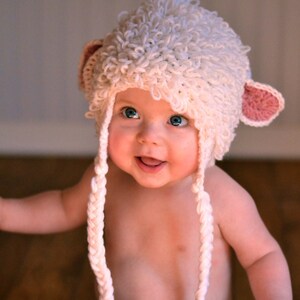 May include: A baby wearing a white crocheted lamb hat with pink ears and blue jeans.