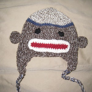 Puede incluir: Un gorro de mono de crochet marrón y blanco con una boca roja y ojos azules. El gorro tiene dos lazos largos que se pueden atar debajo de la barbilla.