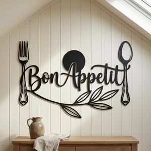 May include: A black metal wall art piece featuring the text Bon Appetit in a cursive script, flanked by a fork on the left and a spoon on the right. A solid black circle sits above the text, and a stylized leafy branch extends below the lettering.