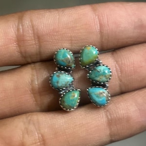 May include: A pair of stud earrings featuring three teardrop-shaped turquoise stones set in silver. Each earring is arranged in a curved, vertical cluster with serrated metal edges surrounding the blue-green stones with brown matrix inclusions.