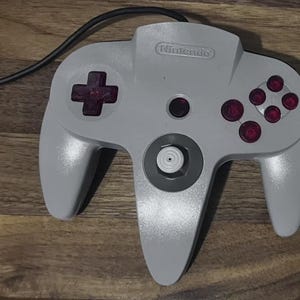 May include: A light grey video game controller with a black cord, featuring a Nintendo logo at the top, a dark purple directional pad, four dark purple action buttons, a central dark purple button, and a white analogue stick with a grey base.
