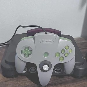 Könnte beinhalten: Ein grauer Nintendo 64-Videospielcontroller mit durchscheinenden lindgrünen Tasten und einem Steuerkreuz, ausgestattet mit einem zentralen Analogstick und dem Nintendo-Logo. Der Controller ist über ein schwarzes Kabel mit einer dunkelgrauen Konsole verbunden.