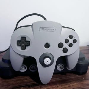 Puede incluir: Un mando de videoconsola Nintendo 64 de color gris con botones negros, una cruceta negra y un stick analógico blanco, apoyado sobre una consola de juegos Nintendo 64 de color gris oscuro. El logo de Nintendo aparece grabado en la parte superior.