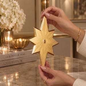May include: A gold-colored, eight-pointed star ornament with a textured, metallic finish. The star features a smooth, flat central surface and a small, raised circular detail in the middle, with decorative, perforated edges along the points.