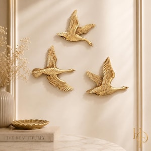 May include: Three gold-colored wall-mounted bird sculptures in flight, each featuring detailed wing textures and outstretched wings. The birds are arranged in a staggered formation, showcasing a metallic finish that reflects light across their surfaces.