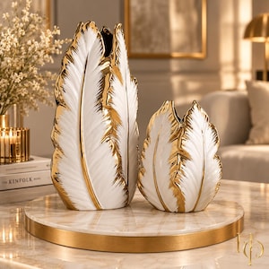 Pode incluir: Dois vasos de cerâmica branca em forma de penas grandes com bordas de ouro metálico. O vaso mais alto está ao lado de um vaso mais curto a combinar. Ambas as peças apresentam um design texturizado com um acabamento brilhante.