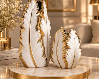May include: Two white ceramic vases shaped like large feathers with metallic gold edges. The taller vase stands next to a shorter, matching vase. Both pieces feature a textured, leaf-like design with a glossy finish and gold-painted details.