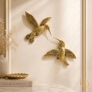 May include: Two gold-colored hummingbird wall sculptures with detailed feather textures. One bird is positioned higher with wings angled upward, while the second bird is positioned lower with wings spread horizontally. Both feature long, pointed beaks.