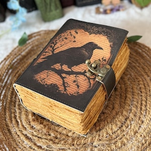 Op de afbeelding: Een rechthoekig dagboek met een donkerbruine kaft met een zwarte kraai-illustratie op een tak tegen een oranje achtergrond. Het boek heeft dikke, getextureerde, lichtbruine papierranden en een metalen, bronskleurige sluiting.