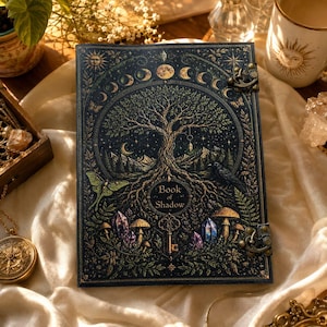 May include: A dark blue hardcover journal titled Book of Shadow featuring a gold foil illustration of a tree, moon phases, a raven, mushrooms, crystals, and a moth. The journal is secured with two antique brass metal latches on the side.
