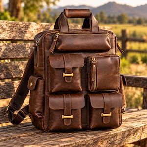 May include: A dark brown leather backpack featuring a top handle, a horizontal zipped compartment, and four front pockets with flap closures secured by gold-toned metal buckles. The bag includes side pockets and adjustable shoulder straps.