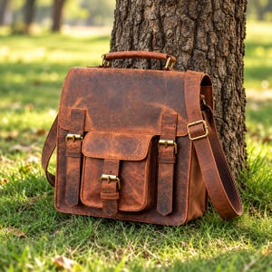 Könnte beinhalten: Eine braune Ledertasche mit Used-Optik, ausgestattet mit einem Tragegriff, einem verstellbaren Schulterriemen und einer Vordertasche. Die Tasche wird mit drei messingfarbenen Metallschnallen und passenden Beschlägen in rechteckiger Form verschlossen.