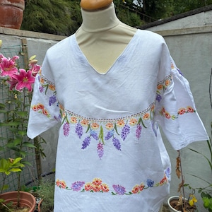 May include: A white short-sleeved blouse featuring a V-neckline and embroidered floral patterns in shades of purple, orange, and green. The embroidery is arranged in horizontal bands across the chest and sleeves, showcasing detailed flower motifs.