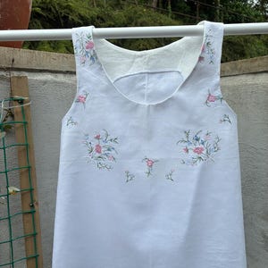 May include: A white sleeveless top featuring delicate floral embroidery in shades of pink and blue. The embroidery is positioned around the scoop neckline and across the centre of the chest, showcasing small flower clusters and stems.