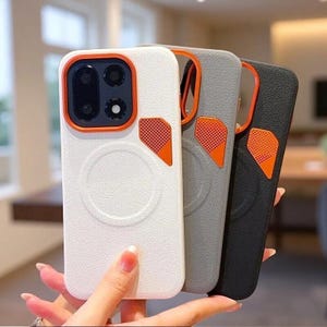 May include: Three smartphone cases in white, grey, and black with a textured finish. Each case features a circular magnetic ring on the back, an orange camera lens frame, and a small orange triangular accent piece with a perforated dot pattern.