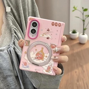 以下が含まれることがあります： 背面に円形の金属製リングスタンドが付いたライトピンクのスマートフォンケース。ケースには、雲の上の小さなキャラクター、星、切手モチーフの漫画風イラストが描かれています。ピンクのフレームがデュアルカメラレンズを囲んでいます。