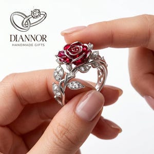 Può includere: Un anello in argento con un design centrale a forma di rosa rosso intenso, accentuato da piccole pietre chiare. La fascia è realizzata per assomigliare a viticci intrecciati con foglie, ognuna incastonata con minuscole pietre chiare.