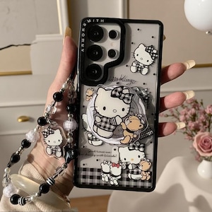 May include: A clear smartphone case with a black frame and camera border labeled ARMOR SMITH. The case features Hello Kitty graphics, a matching circular phone grip, and a beaded wrist strap with black, white, and clear beads and a Hello Kitty charm.