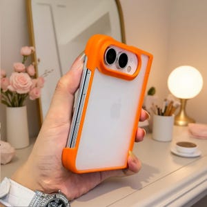 Peut inclure: Une coque de smartphone dotée d'un cadre pare-chocs orange vif et d'un panneau arrière translucide dépoli. La coque comprend une section découpée orange surélevée qui entoure le système de caméra à double objectif du téléphone.