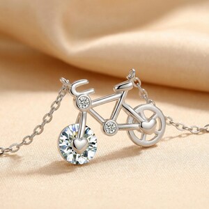 May include: A silver-toned bicycle pendant necklace featuring a large, round, faceted clear crystal as the front wheel, accented with a small heart shape. Two smaller round clear crystals are set in the frame, with a heart on the rear wheel.