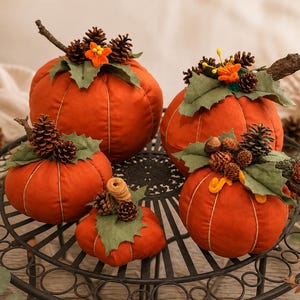 May include: A set of four orange fabric pumpkins of varying sizes, each topped with green felt leaves, small brown pinecones, and orange floral accents. The pumpkins feature stitched vertical lines and are arranged on a black metal table.
